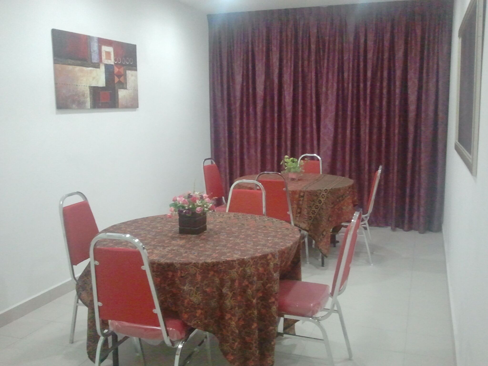 Un coin repas avec une table ronde recouverte d’une nappe à motifs floraux, entourée de chaises rouges. Un pot de fleurs est posé sur la table.