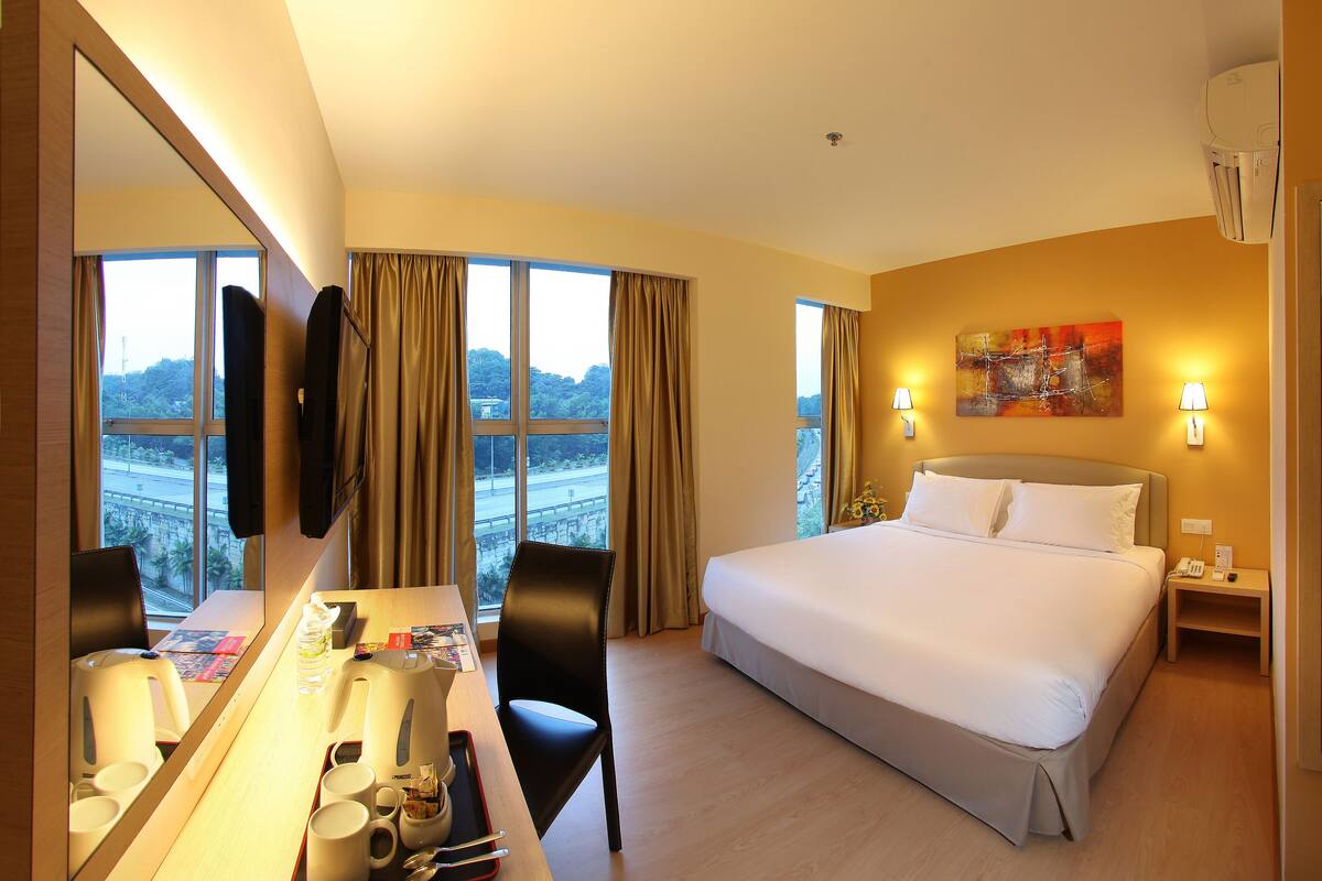 A hotel room with a large bed, a desk with a chair, a mirror, and a view of a bridge and trees.