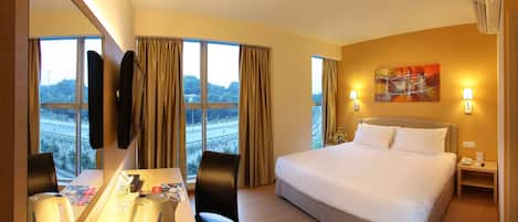 Habitación de hotel con una cama grande, un escritorio con silla, un espejo y vistas a un puente y árboles.