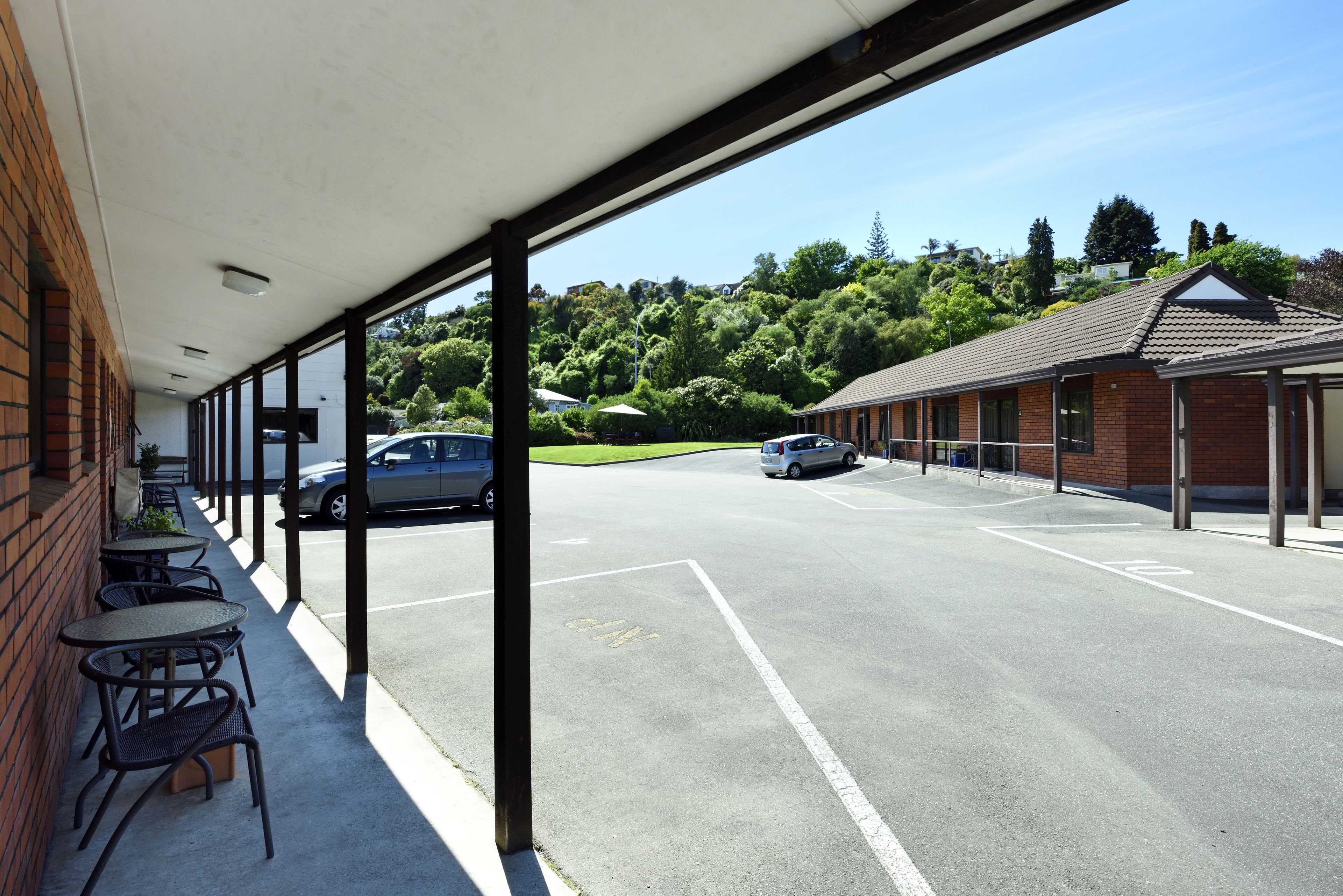 Un espacio al aire libre techado con mesas y sillas, un estacionamiento y un edificio con techo de tejas.