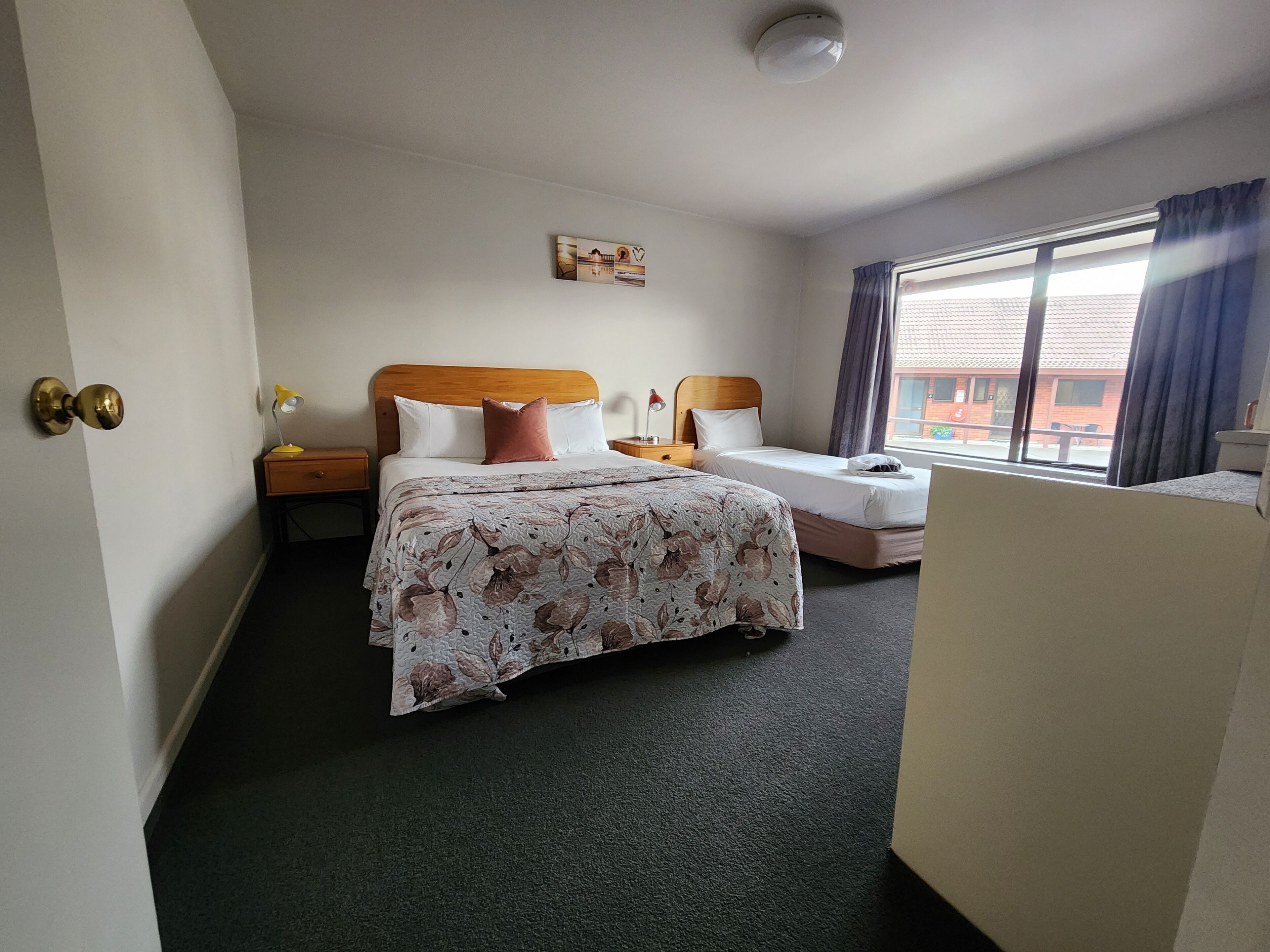 Camera d'albergo con due letti, un'ampia finestra con tende, una lampada a soffitto e un comodino con lampada.