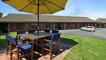 A patio with a wooden table and chairs, a yellow umbrella, and a car parked in front of a building with a brick facade.