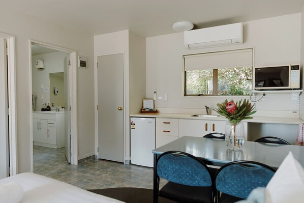 A modern kitchen with white cabinets, a built-in microwave, and a small refrigerator. There is a dining table with blue chairs and a vase with flowers. - Amber Court Motel Nelson (Nelson)