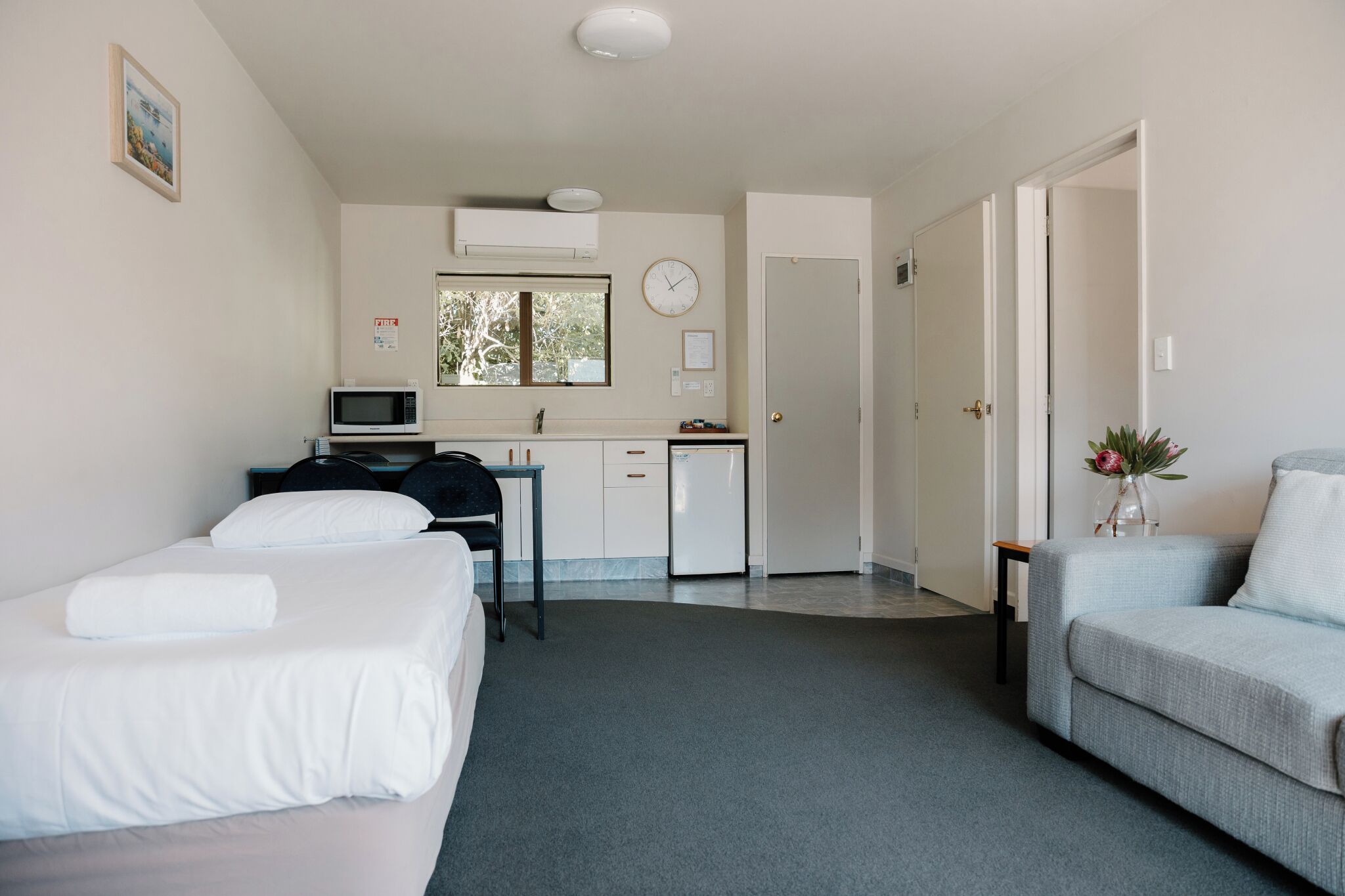 A compact hotel room with a bed, a desk with a computer, a small kitchenette, and a seating area with a sofa.