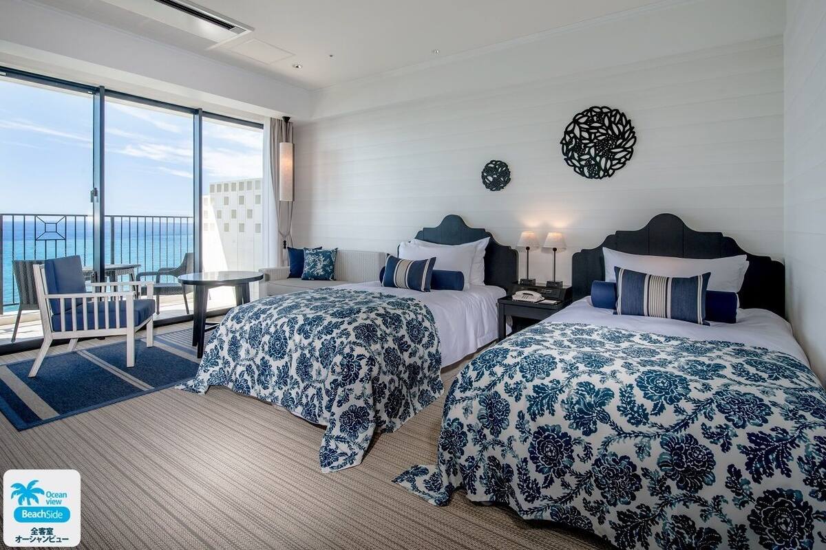 Een hotelkamer met twee bedden, een balkon met uitzicht op zee en een zithoek met een tafel en stoelen.