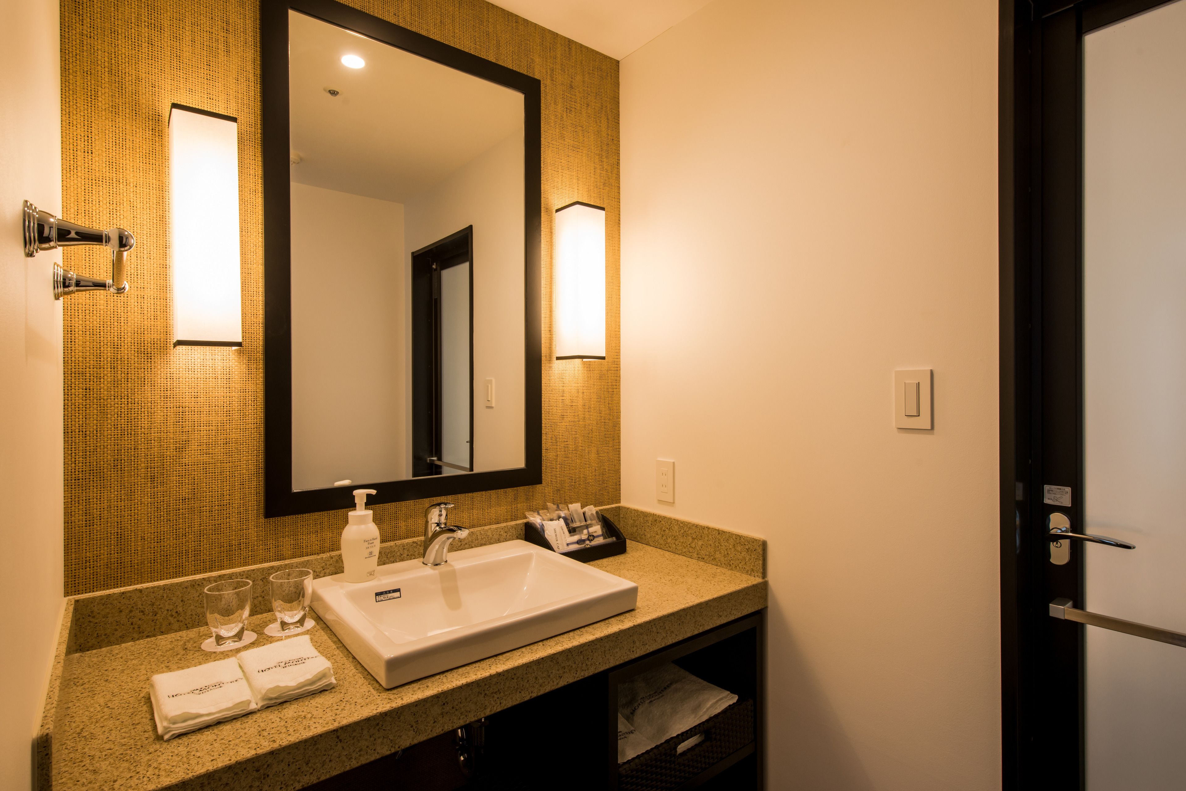 A hotel bathroom with a large mirror, a sink, two glasses, a bottle of soap, and a towel.