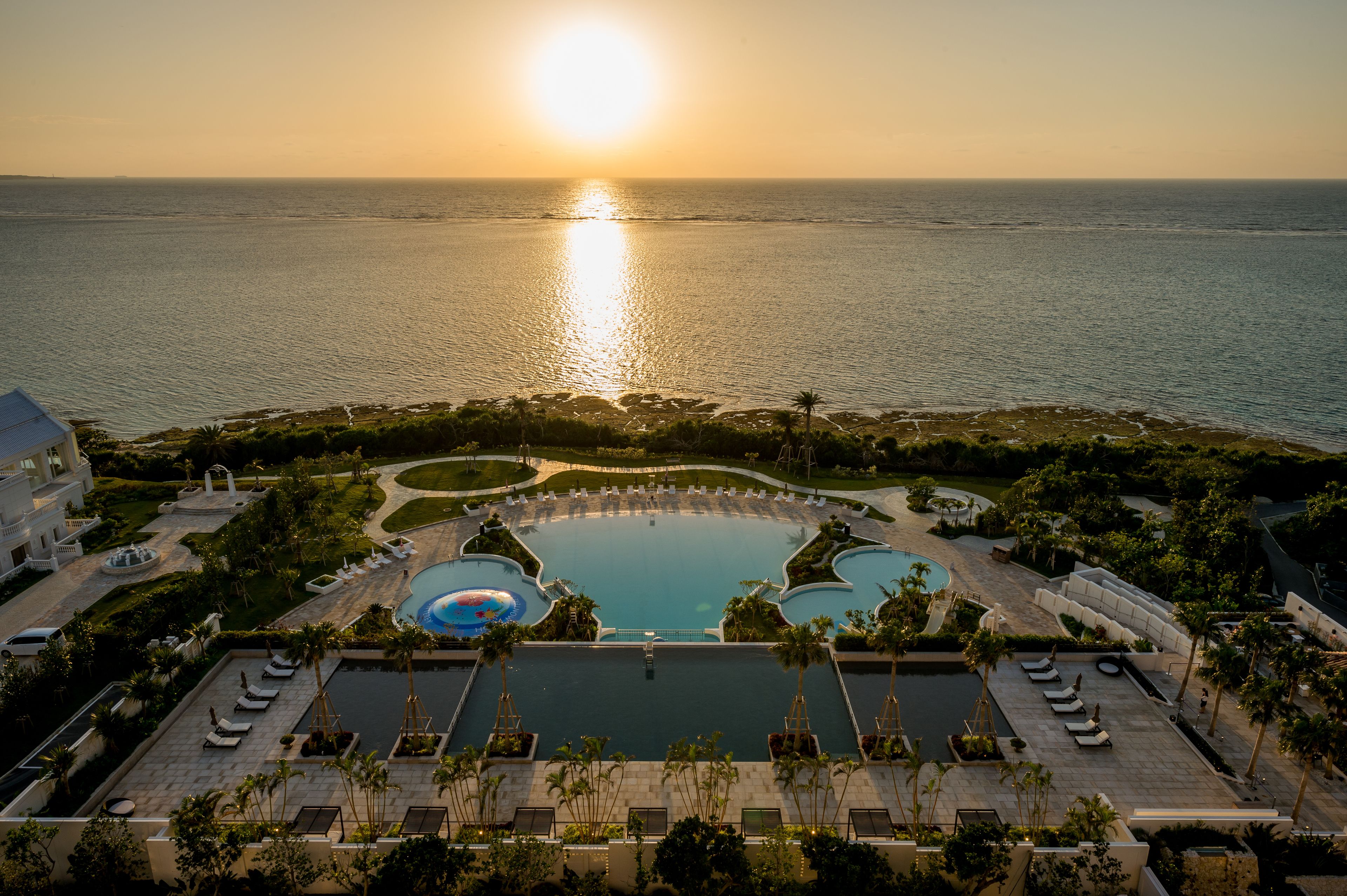 A resort with a large pool, sunbathing areas, and a view of the ocean at sunset.