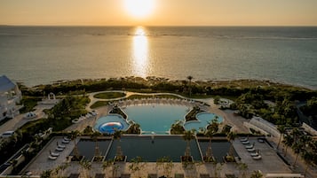 A resort with a large pool, sunbathing areas, and a view of the ocean at sunset.