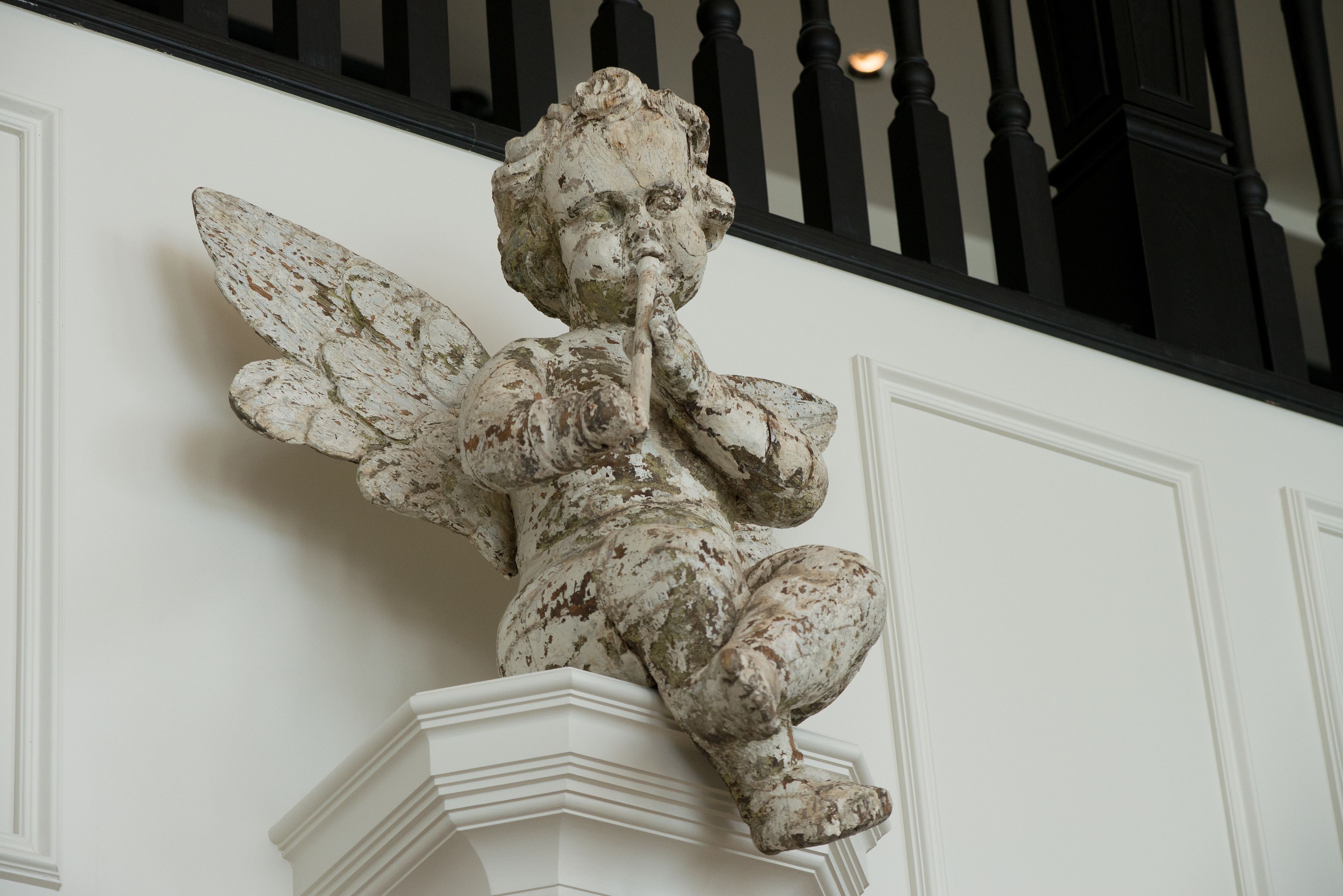 Una estatua blanca y desgastada de un ángel con alas, sobre un pedestal blanco frente a una pared blanca con barandillas negras.
