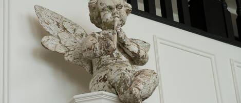 Una estatua blanca y desgastada de un ángel con alas, sobre un pedestal blanco frente a una pared blanca con barandillas negras.