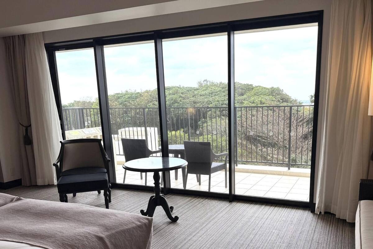 Een hotelkamer met een grote schuifglazen deur die leidt naar een balkon met uitzicht op bomen en een hek. Er staat een bed, een stoel en een kleine tafel met stoelen op het balkon.