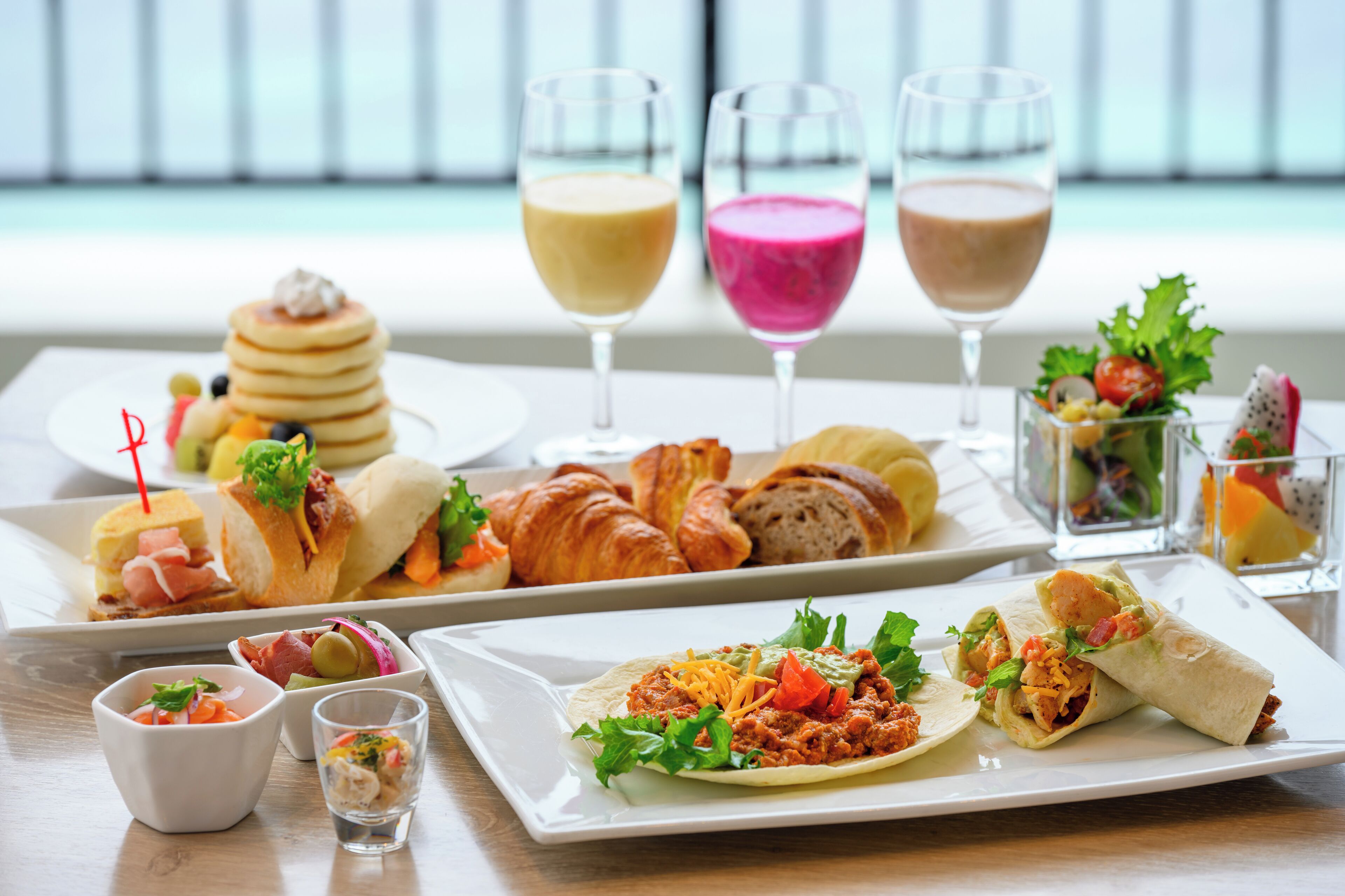 Un bufé de desayuno con diversos platos, entre ellos panqueques, cruasanes y wraps, junto con dos vasos de bebidas.