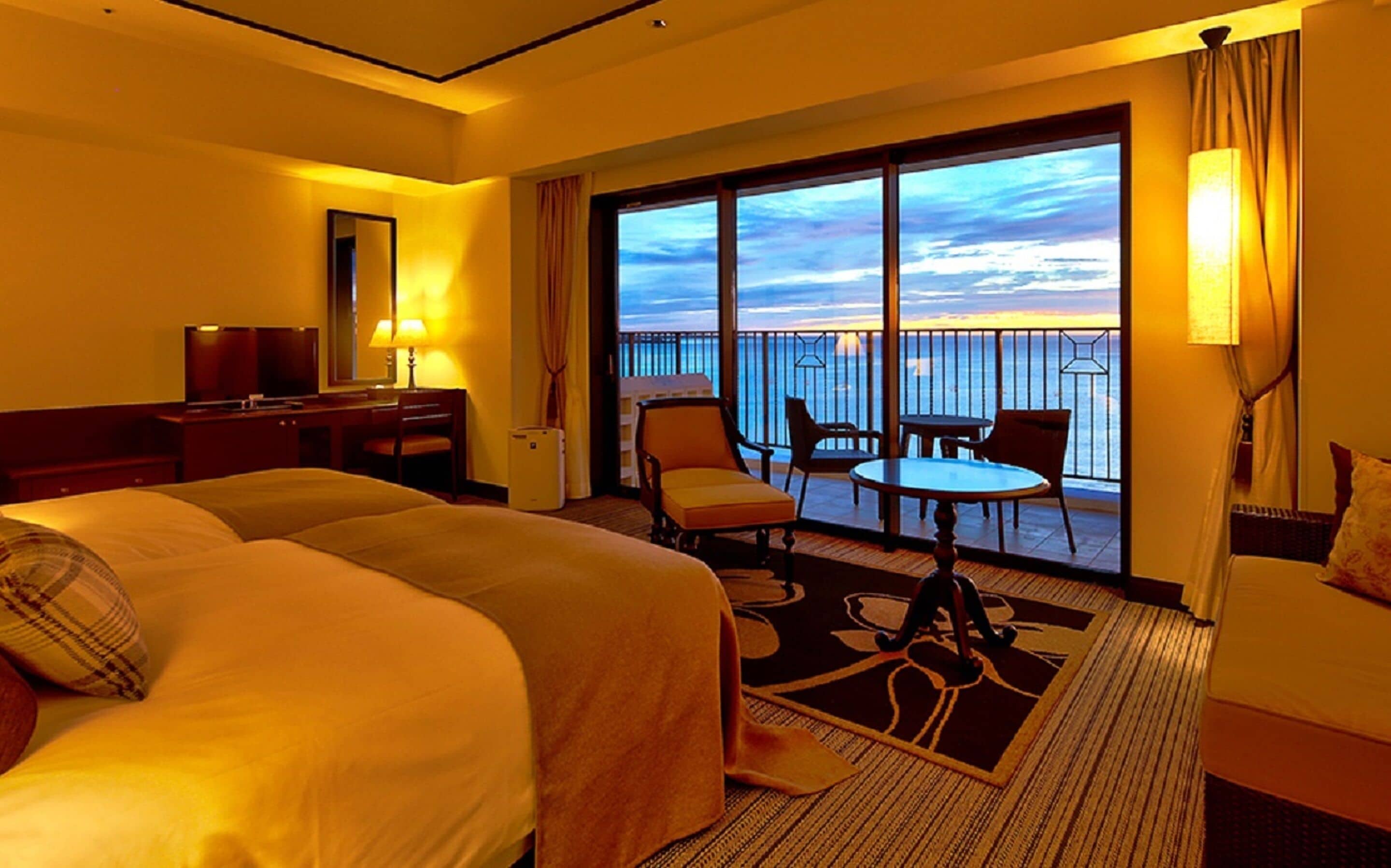 A hotel room with a large bed, a desk, a chair, a small table, and a balcony with a view of the ocean.