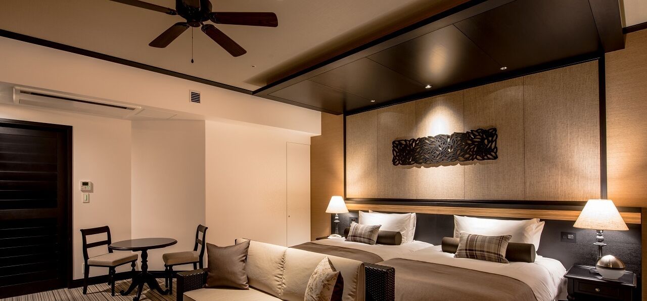 A hotel room with a bed, two bedside lamps, a ceiling fan, a small table with chairs, and a decorative wall piece.