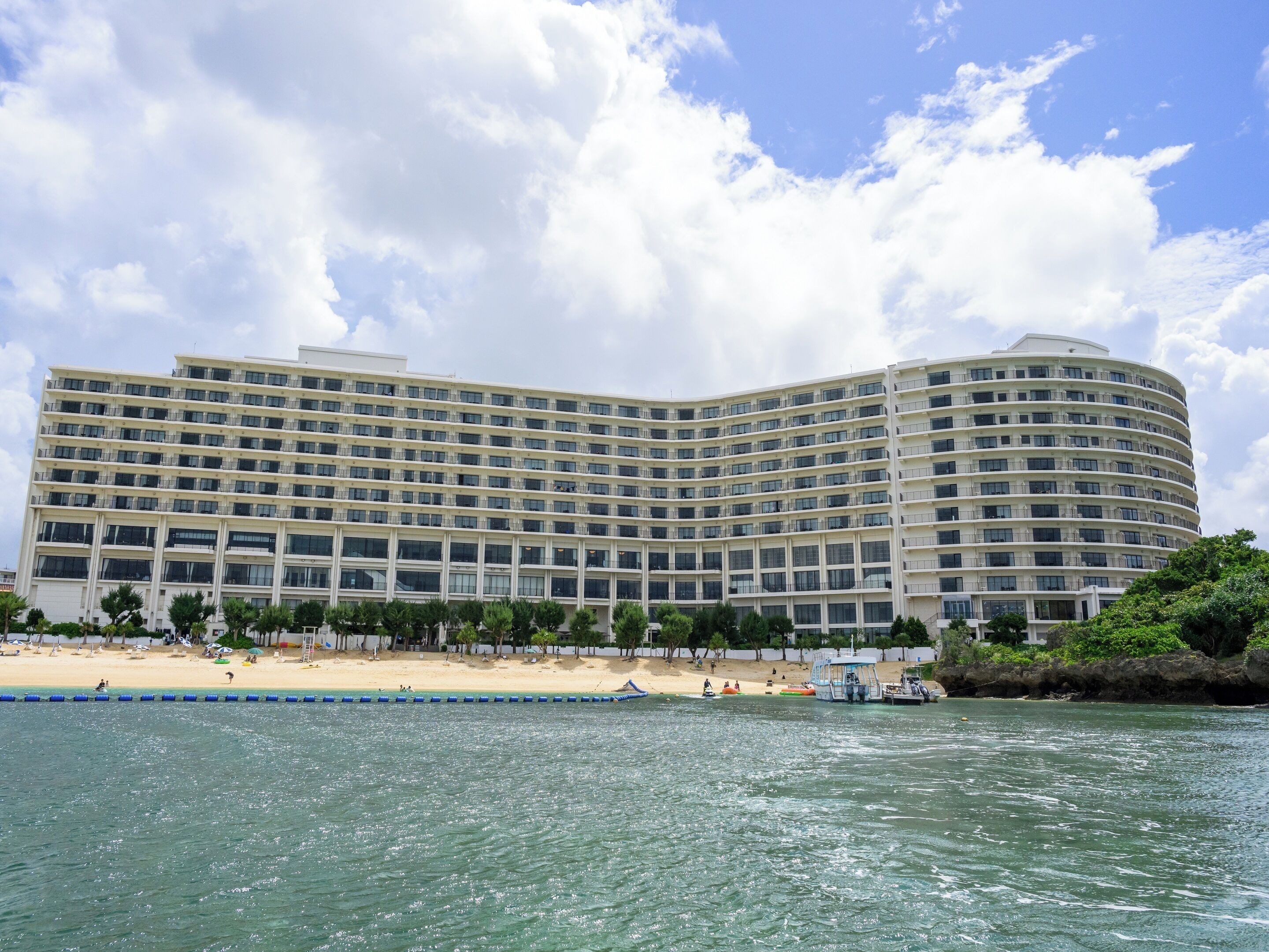A large, multi-story hotel with a curved facade, located near a beach with a swimming area.