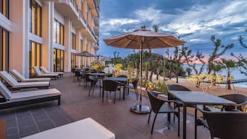A hotel patio with lounge chairs, tables, and an umbrella, overlooking a beach and ocean.