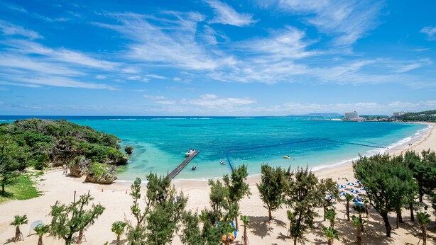 Uma praia com águas turquesa cristalinas, um cais e um hotel ao fundo.