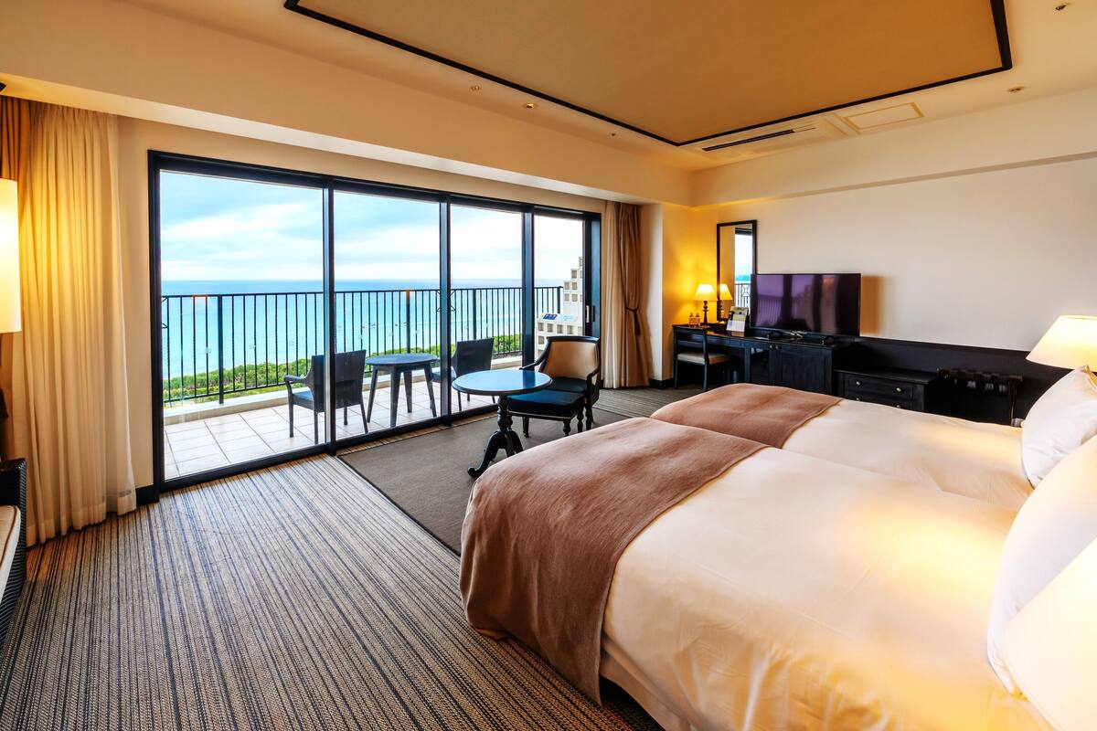 Een hotelkamer met een groot bed, een bureau met een televisie, een balkon met uitzicht op zee, en een eethoek met stoelen.