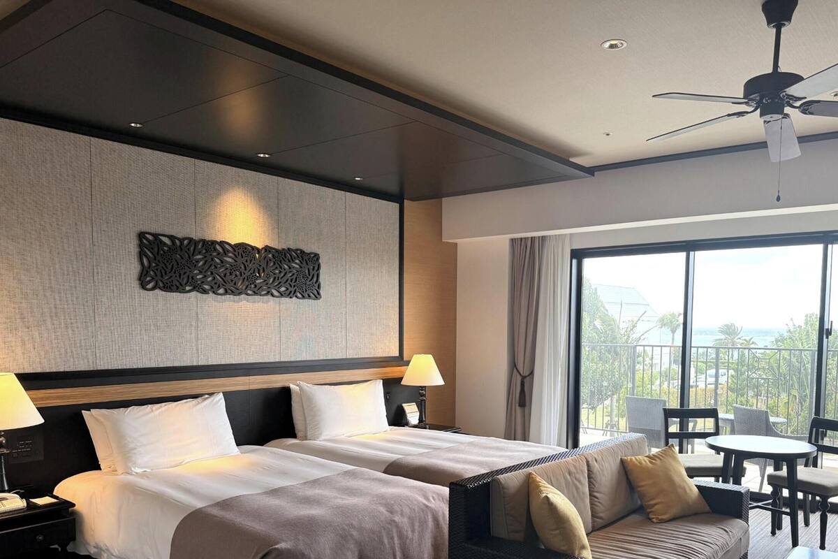 Hotelkamer met twee bedden, een plafondventilator, een balkon met tuinmeubilair en een decoratief wandobject.
