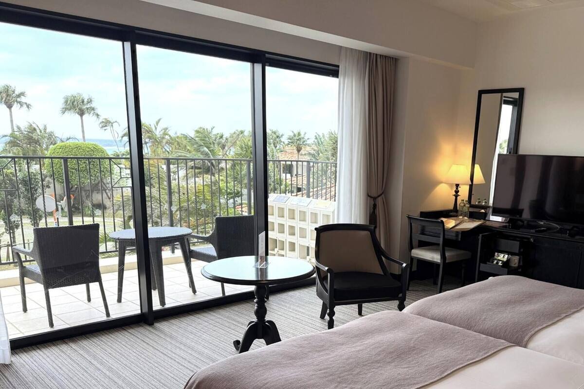 Een hotelkamer met een groot balkon, een bed, een bureau met een televisie en een stoel.