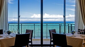 Uma sala de jantar com uma mesa posta para dois, com vista para um mar azul-turquesa e céu claro.
