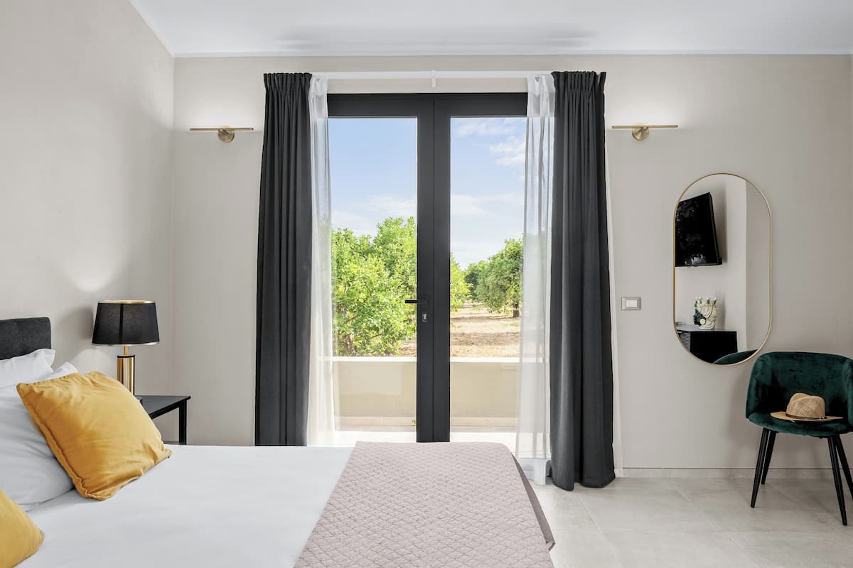 Habitación de hotel con cama, mesita de noche, lámpara, silla, espejo y vistas al exterior con vegetación.