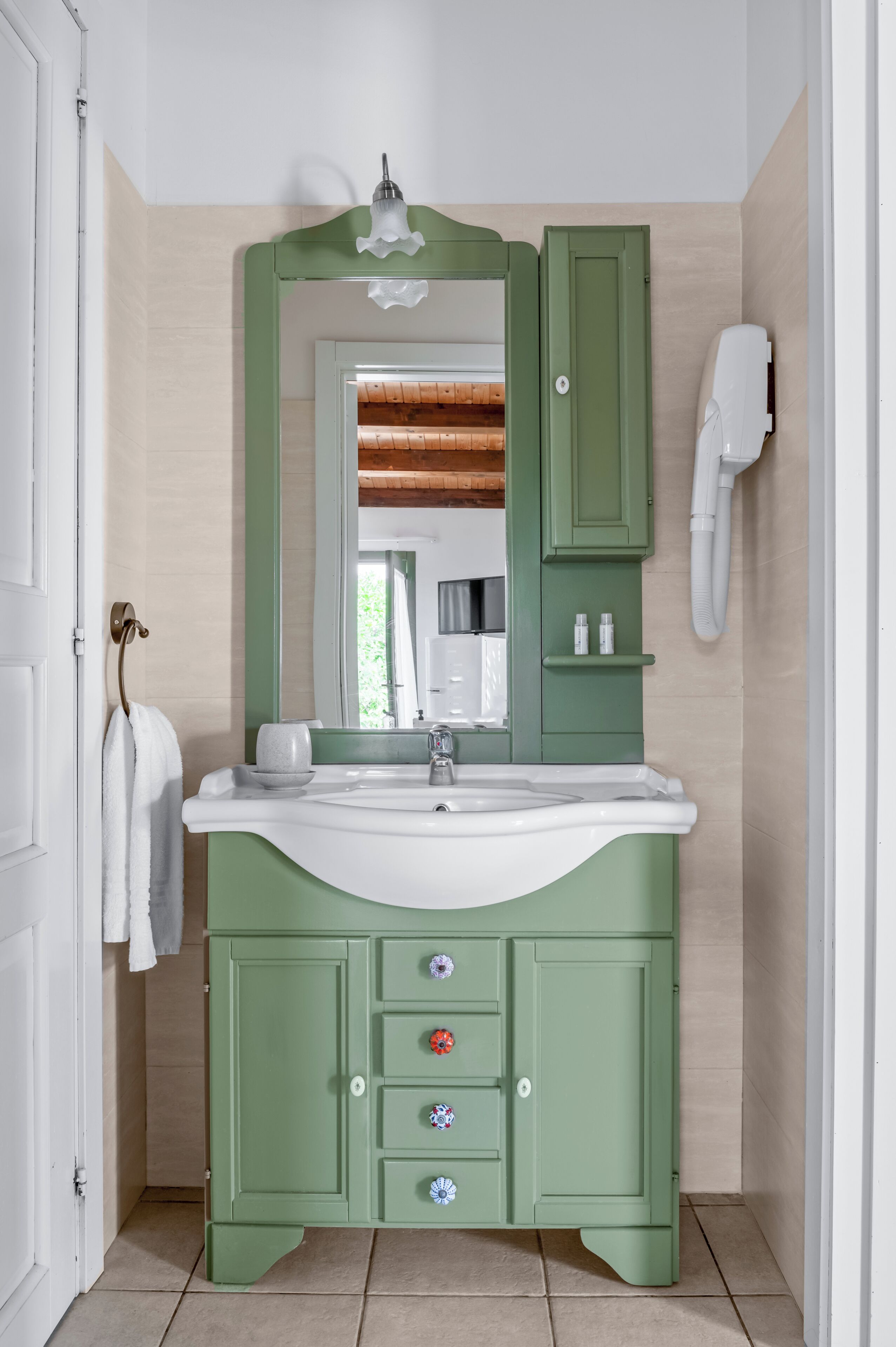 A bathroom with a green vanity, a white sink, and a mirror. There is a towel hanging on the door and a hairdryer on the wall.