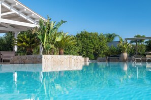 Piscina azul cristalina con paredes de piedra, rodeada de exuberante vegetación y una pérgola blanca.