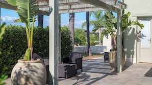 Een terras buiten met een pergola, zitplaatsen en grote potplanten.