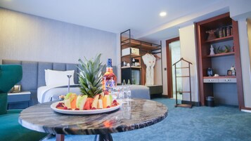 A hotel room with a bed, a green armchair, a round table with fruit and a bottle, and a wardrobe with shelves.