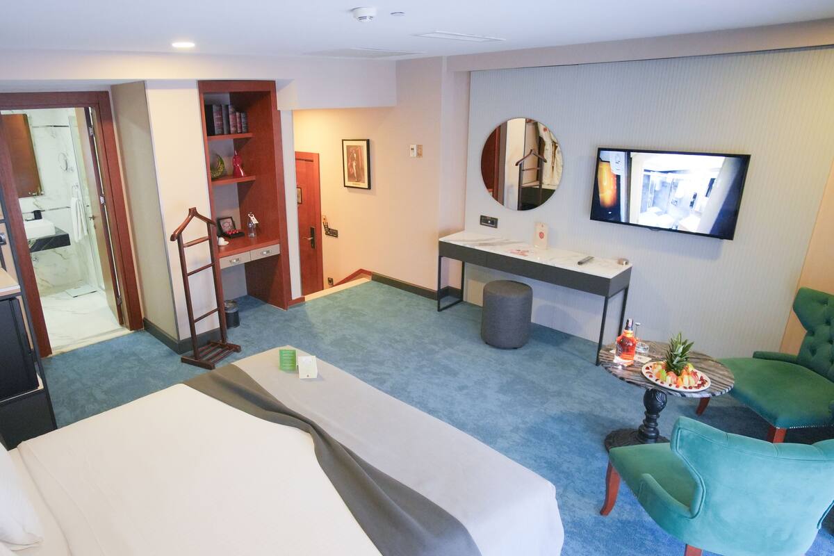 A hotel room with a bed, a desk with a mirror, a television, a small table with fruit, and a green armchair.