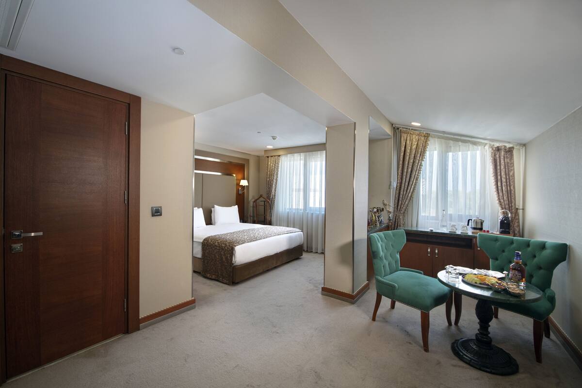 A hotel room with a bed, a dining table with chairs, a minibar, and a view of the outside through the window.