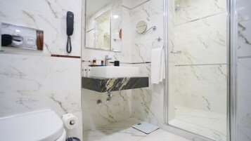 Un baño moderno con piso de mármol, cabina de ducha de vidrio, lavamanos blanco y teléfono fijo en la pared.