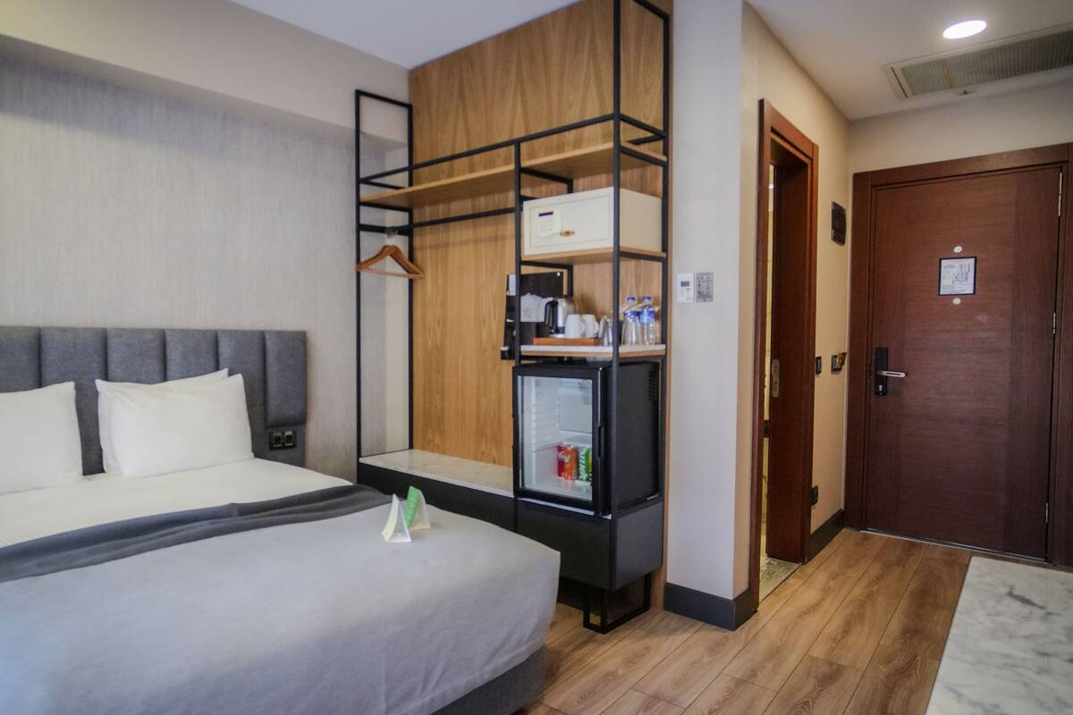 A hotel room with a bed, a bedside table, a small refrigerator, a wooden shelf unit, and a door.
