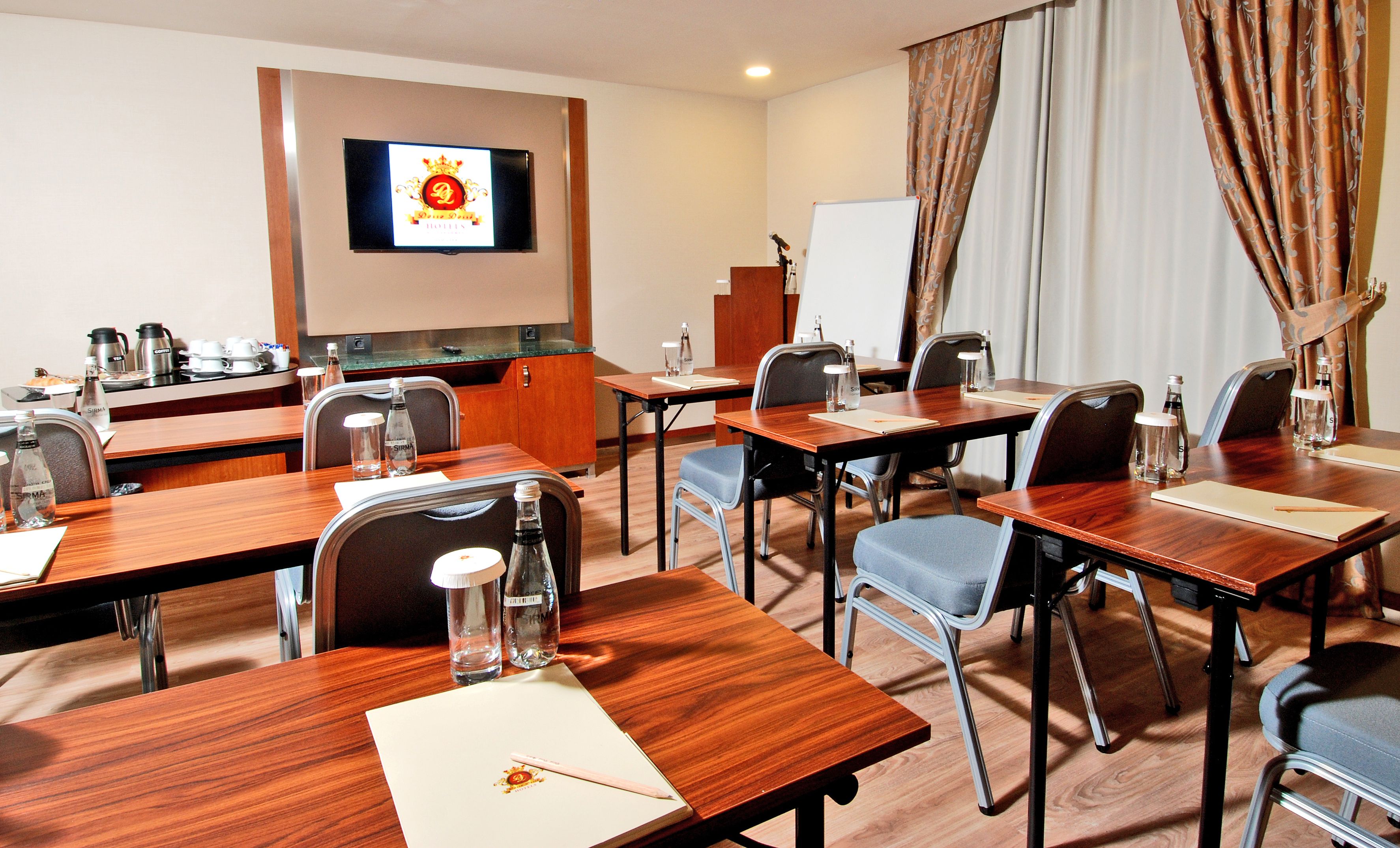 Un salón de conferencias de hotel con mesas y sillas de madera, una pizarra, una pantalla de televisión y un dispensador de agua.