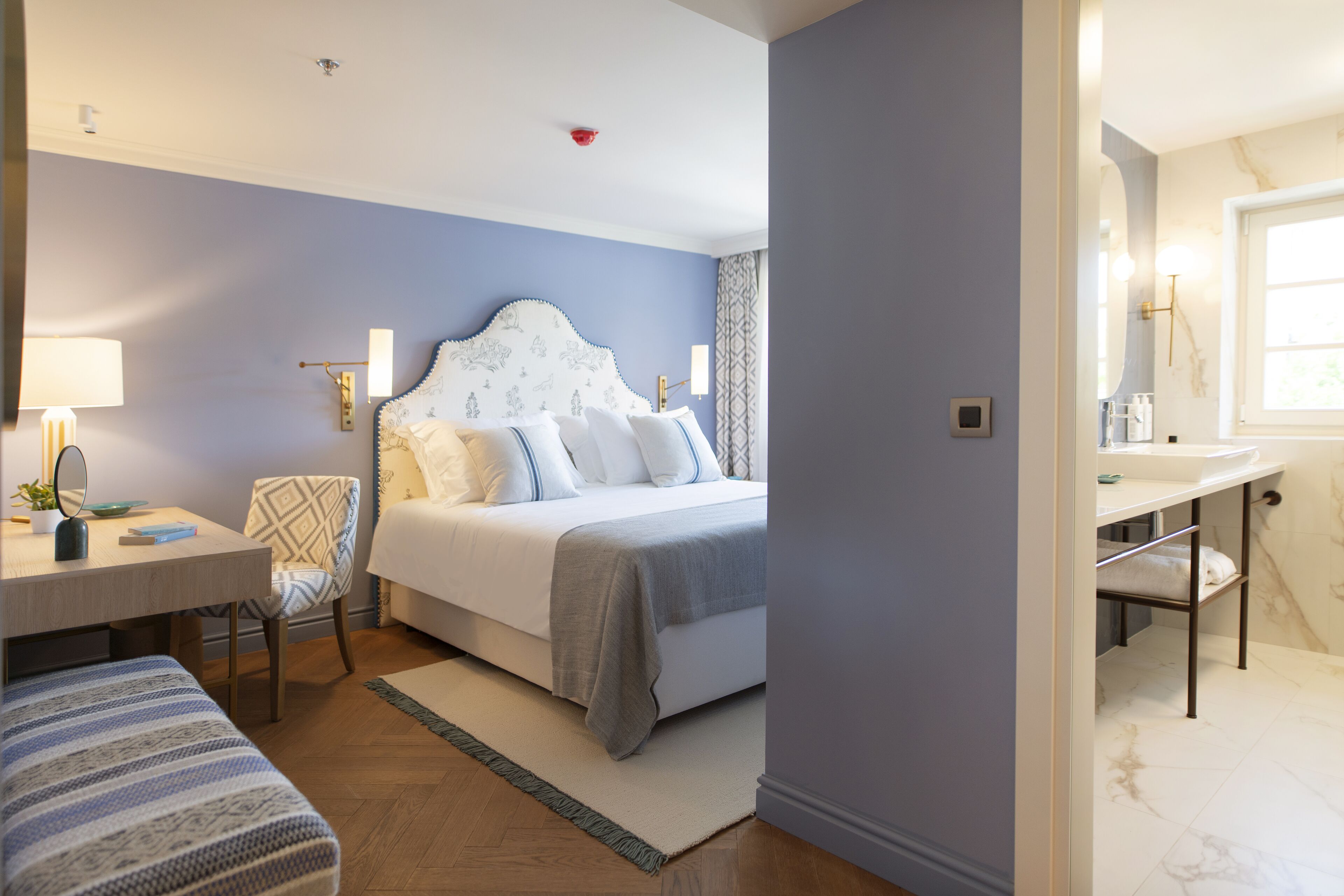 A bedroom with a bed, bedside table, chair, and a small desk with a lamp.