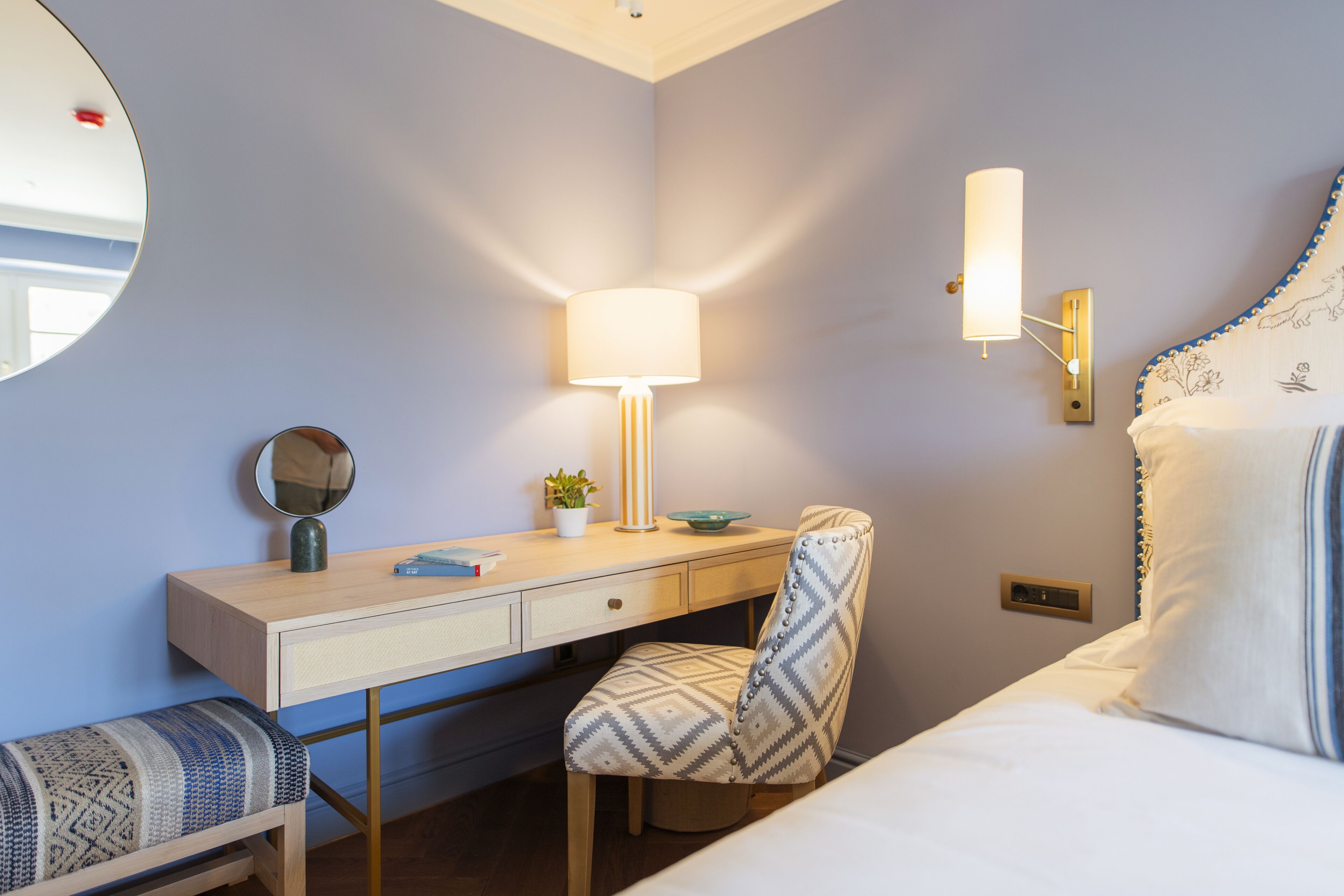 Habitación de hotel con cama, escritorio con lámpara, una silla, espejo y lámpara de pared.