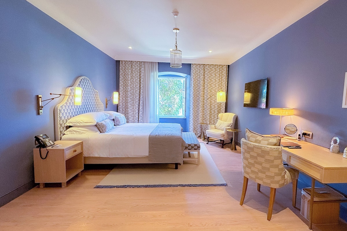 A hotel room with a large bed, bedside tables, a desk, a chair, a window with curtains, a ceiling light, and a wall-mounted TV.
