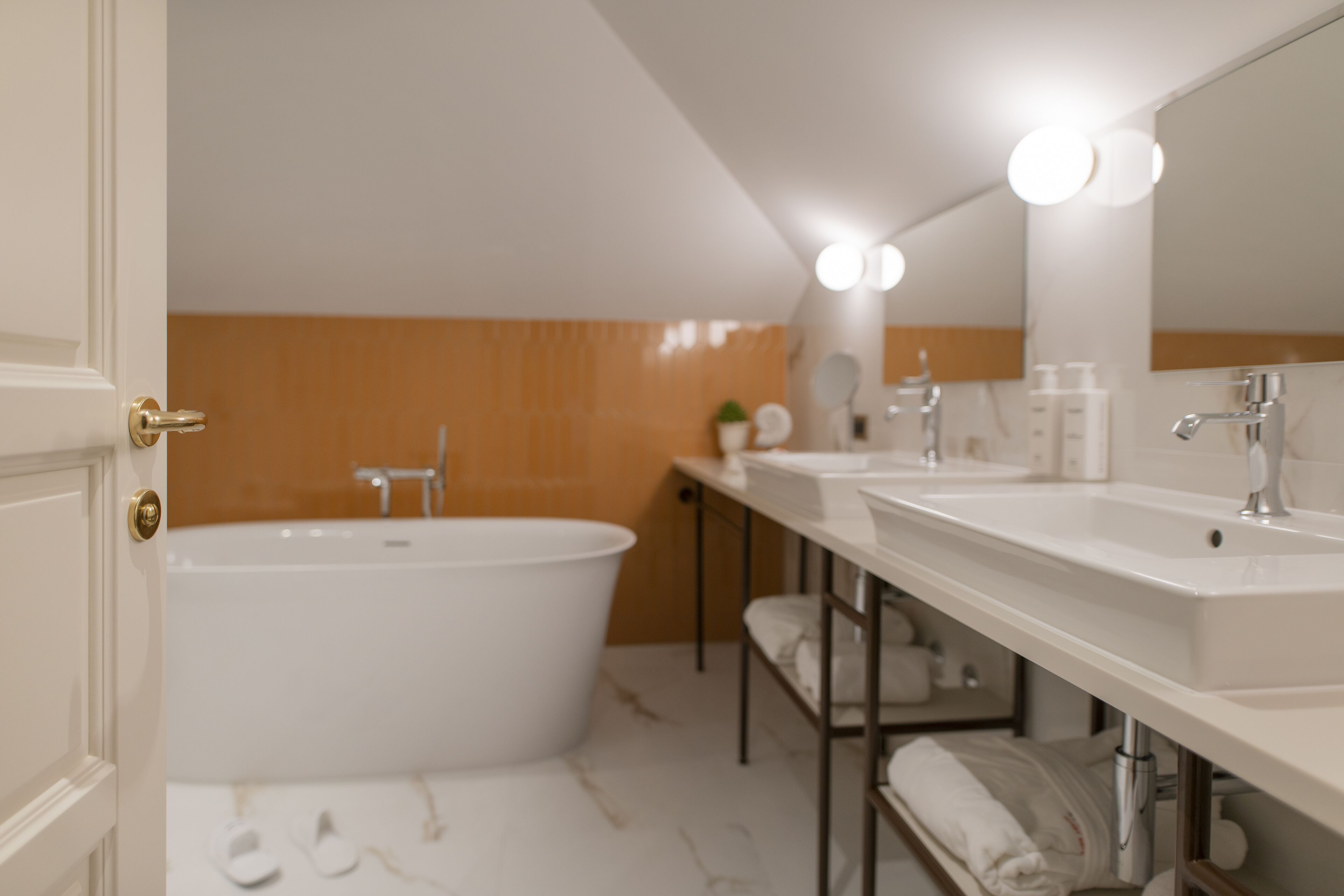 A modern bathroom with a bathtub, two sinks, and a large mirror with lights.