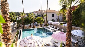 A pool area with lounge chairs and umbrellas, surrounded by palm trees and a building with a tiled roof. - Hotel Supetar Cavtat (Konavle)