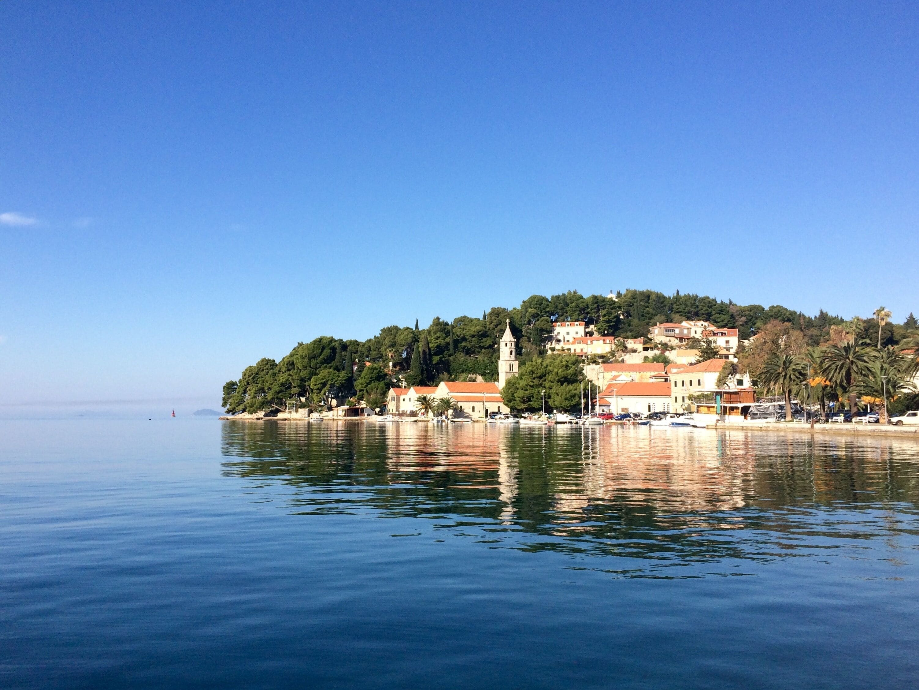 A coastal town with buildings on a hillside, a church with a bell tower, and calm water reflecting the structures.