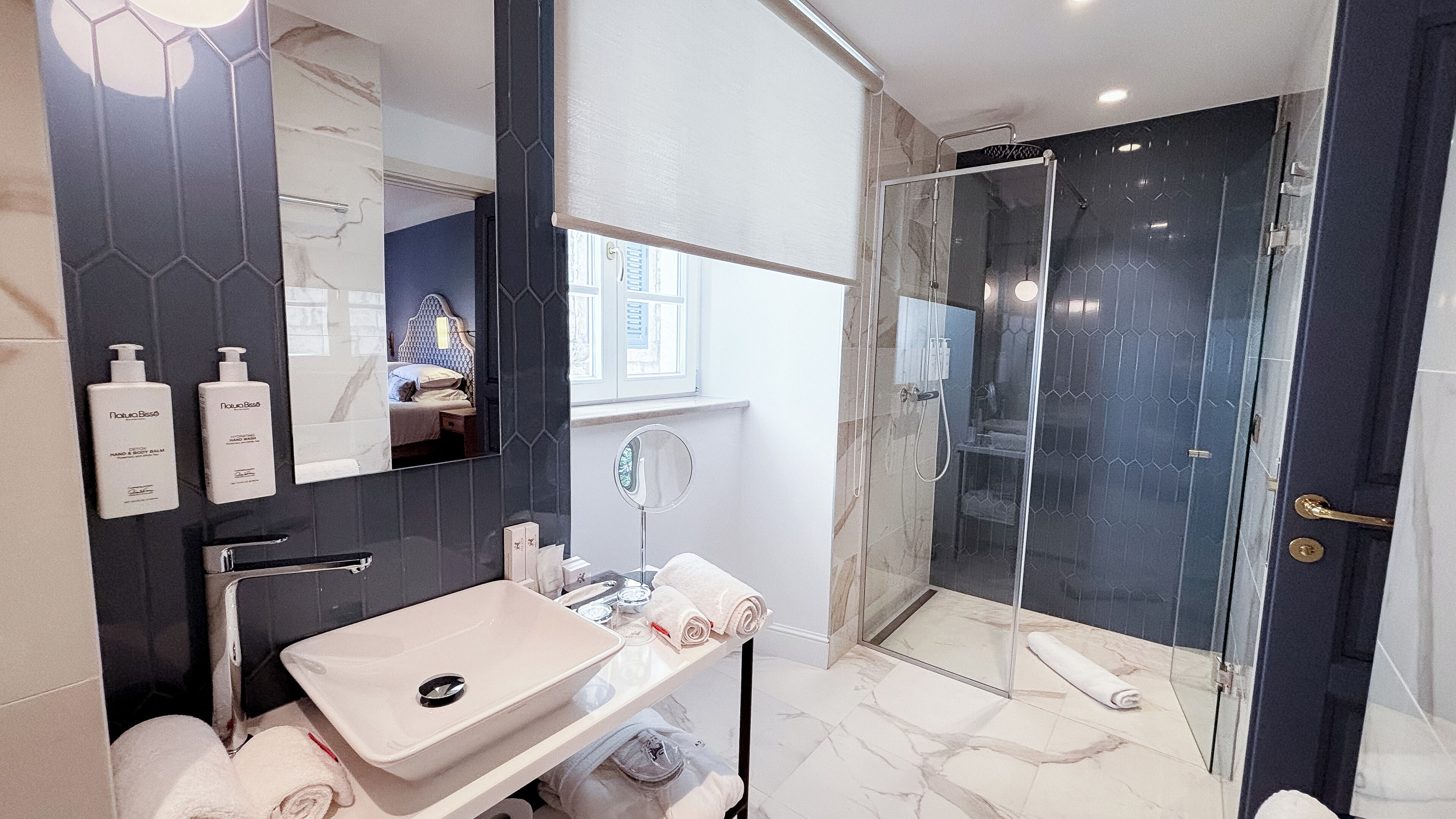 A modern bathroom with a large mirror, a sink, a glass-enclosed shower, and toiletries on the wall.