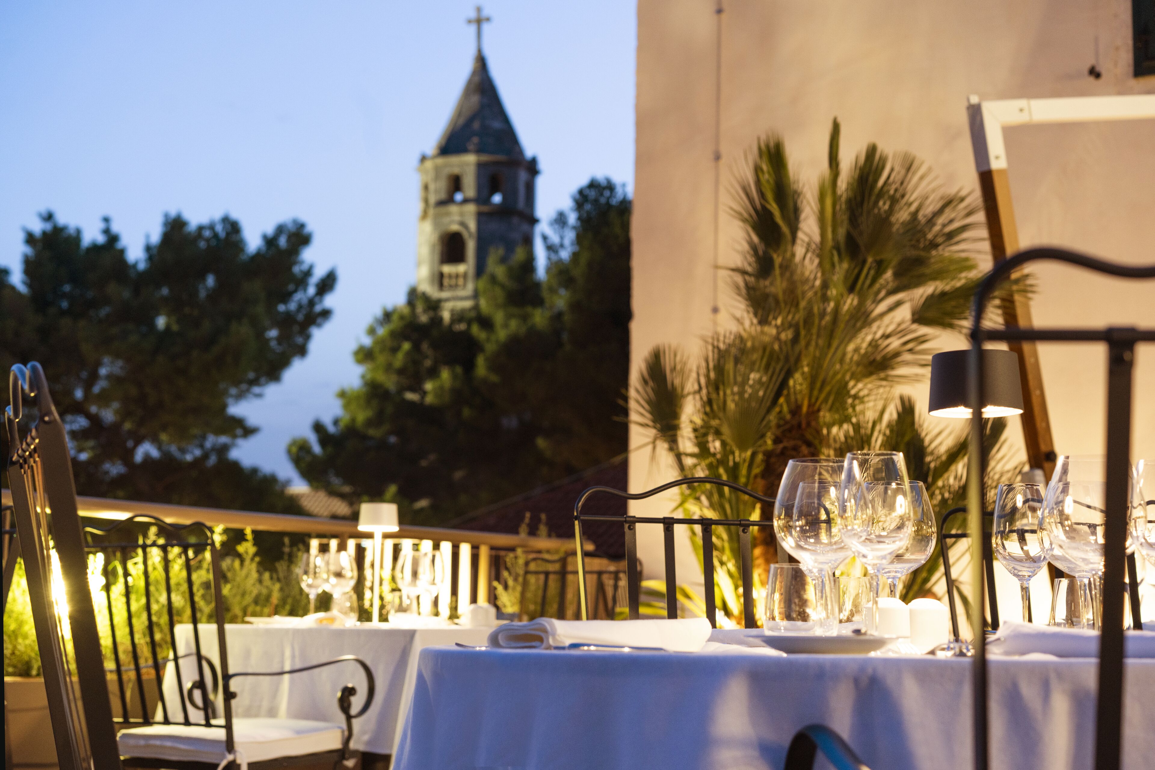 Un área de comedor al aire libre con mesas dispuestas para una comida, una torre de iglesia al fondo y palmeras.