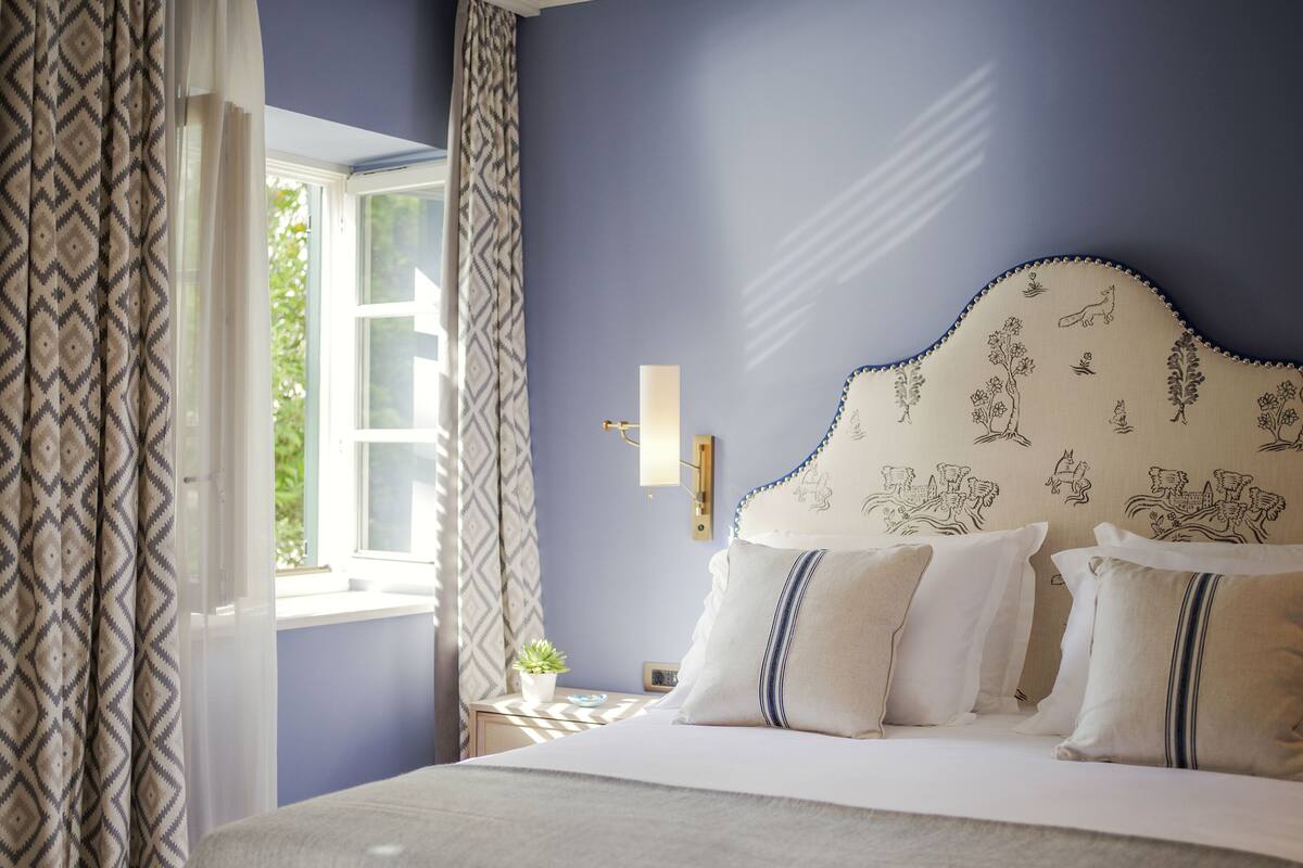 A bedroom with a bed, patterned curtains, a wall-mounted light, and a window with a view of greenery.