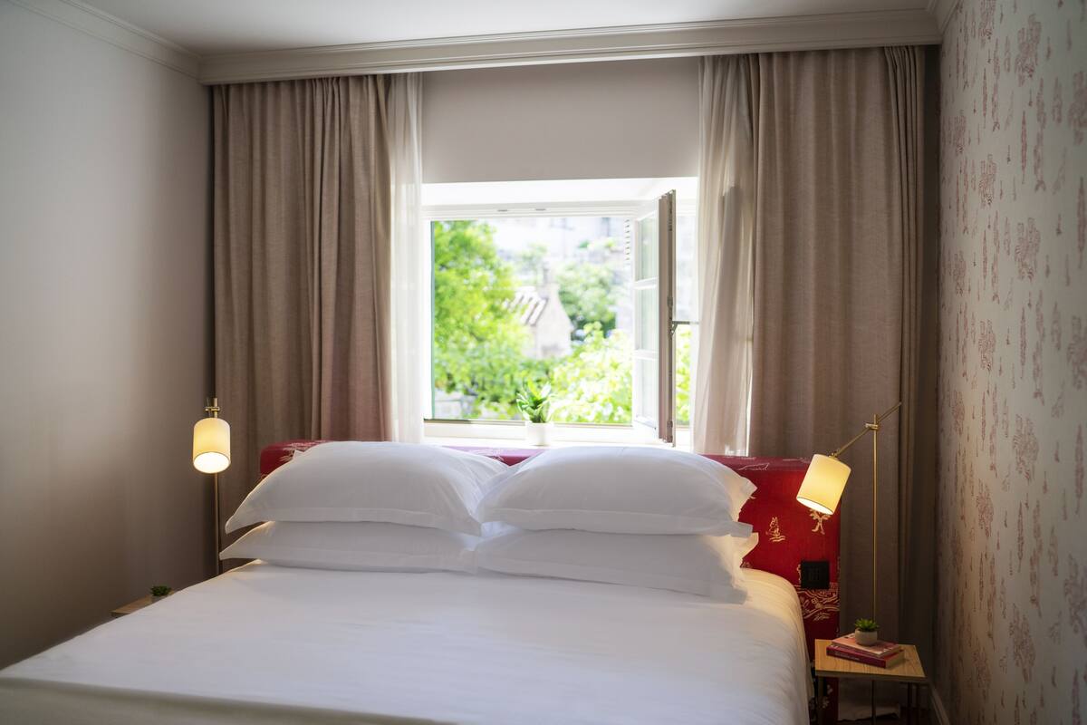 A hotel room with a bed, curtains, a window with a view of greenery, a lamp, and a small table with a plant.