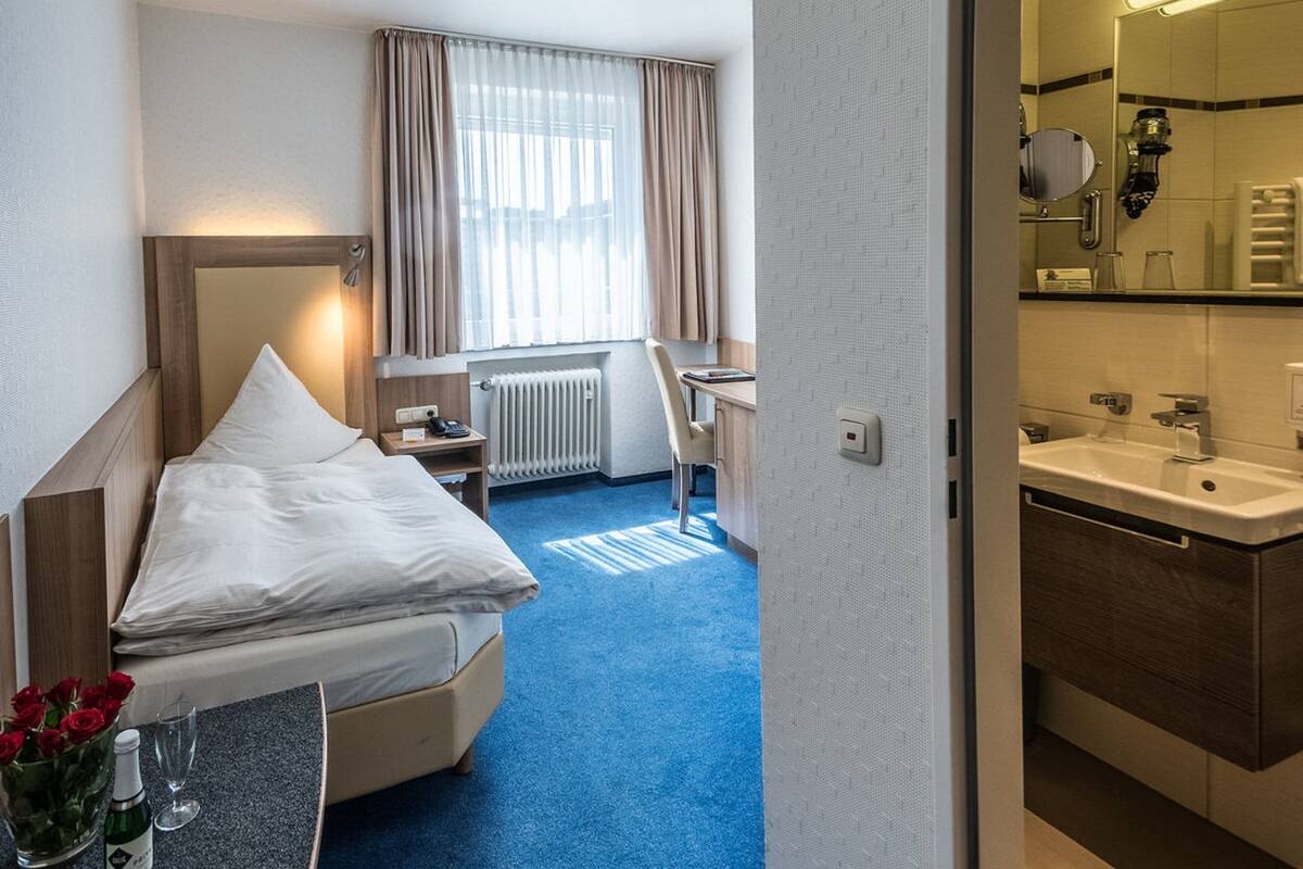 Une chambre d’hôtel avec un lit, un bureau, une chaise, une petite table ornée d’un vase de fleurs et une salle de bain visible à travers une porte ouverte.