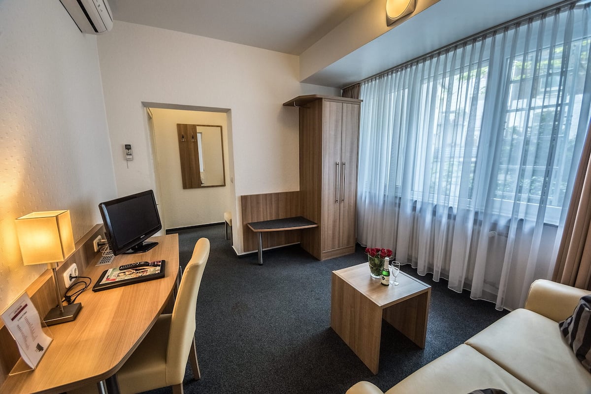 Une chambre d’hôtel équipée d’un bureau, d’une chaise, d’une petite table ornée d’un vase de fleurs, d’une armoire et d’une grande fenêtre avec des rideaux.