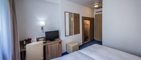 A hotel room with a bed, a desk with a computer, a chair, a TV, a mirror, and a wardrobe.