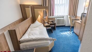 Una camera d'albergo con un letto, una scrivania, una sedia, un tavolino con un bicchiere e una bottiglia, e una finestra con tende.