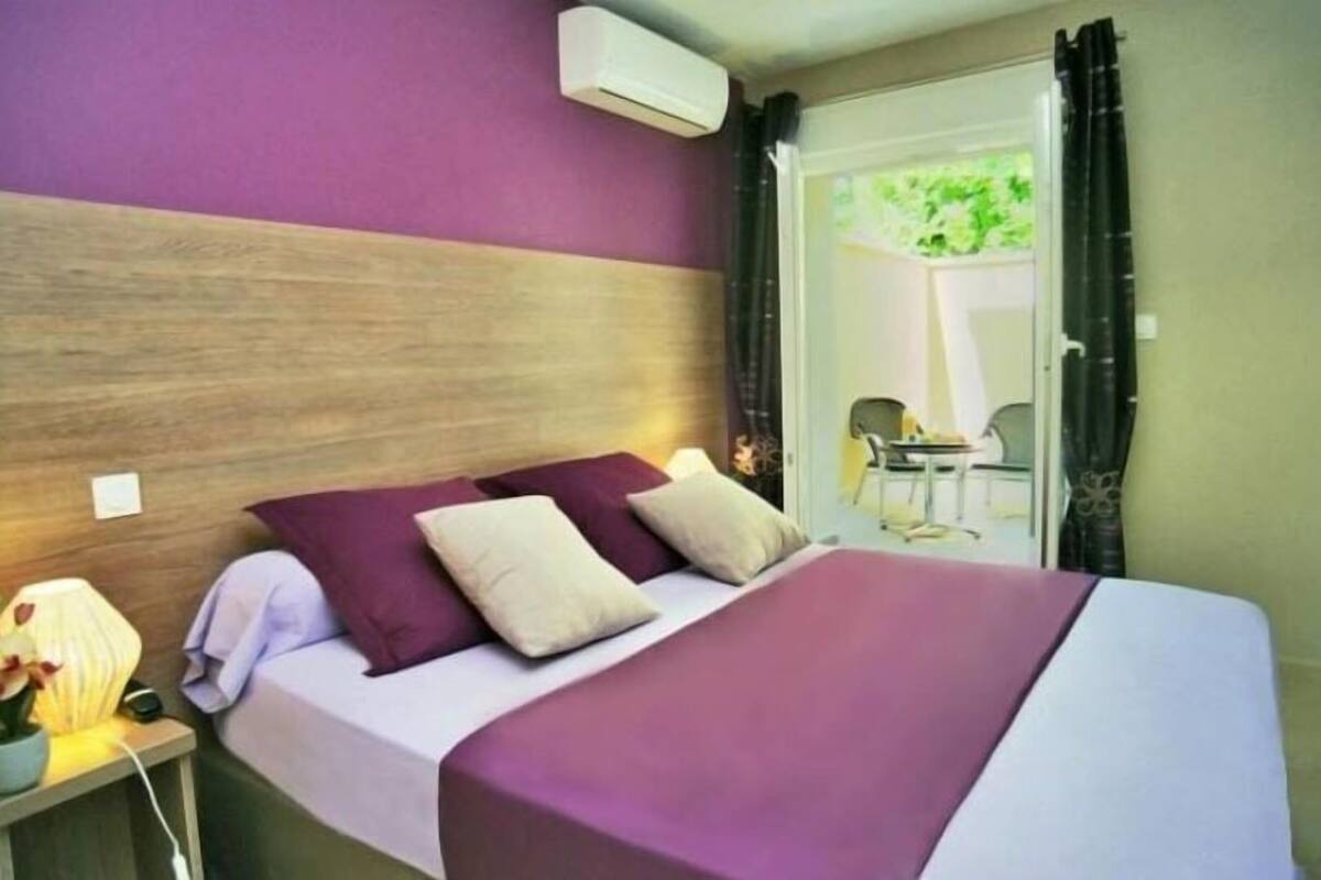 A bedroom with a wooden headboard, a bed with a purple and white bedspread, and a nightstand with a lamp and a vase of flowers.